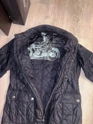 Svart quiltad jacka från Barbour - Snygg svart quiltad jacka från Barbour med hög krage, tryckknappar och dragkedja. Jackan har två framfickor med lock och ett coolt motorcykeltryck på insidan. Perfekt för dig som gillar klassiskt. Jackan är använd någon enstaka gång bara. Hör av dig vid minsta fundering!🙂