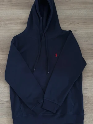 Mörkblå hoodie från Polo Ralph Lauren - Snygg mörkblå hoodie från Polo Ralph Lauren med klassisk röd broderad logga på bröstet. Hoodien har huva med dragsko, känguruficka och ribbade muddar. Perfekt för en avslappnad och stilren look.