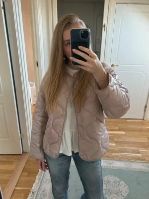 Ljusrosa quiltad cardiganjacka - Säljer en ljusrosa quiltad cardiganjacka med rund hals och knäppning framtill. Jackan har ett vågigt mönster i quiltningen och är lätt vadderad, perfekt för lager-på-lager. Passar till jeans och t-shirt för en avslappnad look.