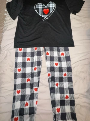 Svart pyjamas med rutiga hjärtbyxor - Mysig pyjamas med svart t-shirt och stort rutigt hjärta med röd detalj på bröstet. Byxorna är vita och svarta i rutigt mönster med röda hjärtan över hela. Perfekt för hemmakvällar och chill. Mjukt material som känns skönt mot huden.