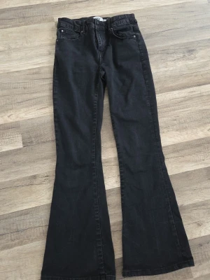 Svarta bootcut jeans med medel högmidja - Säljer ett par svarta bootcut jeans med medelhög midja och klassisk femficksdesign. Jeansen har utsvängda ben och stängs med dragkedja och knapp. Perfekta för dig som gillar en retroinspirerad look.