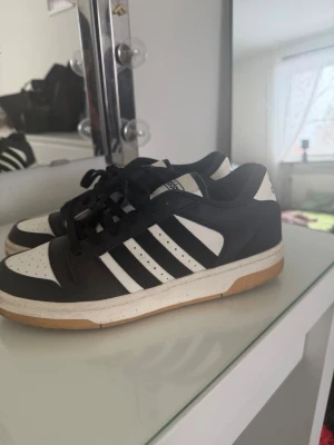 Svarta och vita Adidas sneakers - Säljer ett par klassiska Adidas sneakers i svart och vitt med de ikoniska tre ränderna på sidan. Skorna har svart ovandel i läder, vita detaljer och gummisula i beige. Låga i modellen och med svart snörning. Perfekta för en clean och sportig look. Väldigt sköna och bekväma köpte dem för 800. 