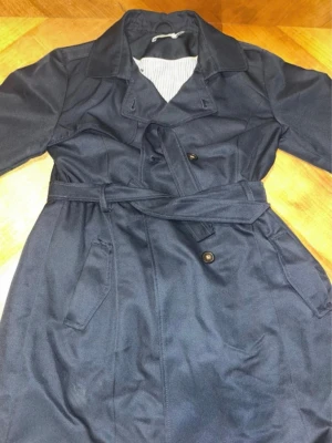 Mörkblå trenchcoat med bälte - Stilren mörkblå trenchcoat med klassisk krage, knappar framtill och matchande bälte i midjan. Jackan har två snedställda fickor och detaljerade manschetter med knapp. Perfekt för dig som vill ha en tidlös och snygg look. Jätte fin och bekväm storlek 158-164