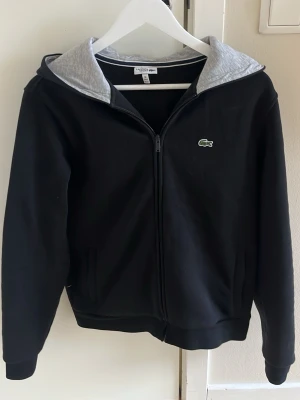 Svart hoodie med dragkedja från Lacoste - Snygg svart hoodie från Lacoste med dragkedja framtill och grå insida på huvan. Klassisk krokodillogga på bröstet och två fickor. Tillverkad i mjukt bomullsmaterial, perfekt för en avslappnad stil. Står att den passar 176 men passar mer runt 170