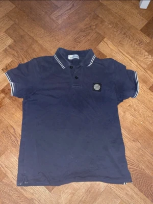 Marinblå pikétröja från Stone Island - Klassisk marinblå pikétröja från Stone Island med korta ärmar och vit kontrastkant på krage och ärmslut. Tröjan har två små hål men det är inget som märks när den är på 