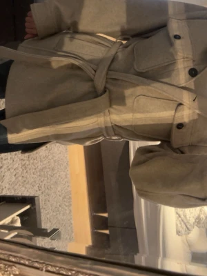 Beige kappa med knytskärp - Stilren beige kappa med knytskärp i midjan och stora fickor framtill. Kappan har knappstängning, klassisk krage och är tillverkad i ett mjukt tyg som ger en avslappnad men trendig look. Perfekt för dig som gillar minimalistisk stil.