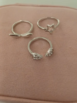 Söta silvriga ringar med stjärna & hjärta - Tre silvriga ringar i olika designer: en med två vingar en med en stjärna och en öppen ring med två hjärtan dekorerade med små glittrande stenar. Perfekta för att mixa och matcha till din stil. Aldrig använt.