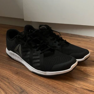 Svarta träningsskor från Reebok - Säljer ett par svarta träningsskor från Reebok med vit sula och meshdetaljer för extra ventilation. Skorna har snörning och en sportig design med Reebok-logga på sidan. Perfekta för gymmet eller löprundan.