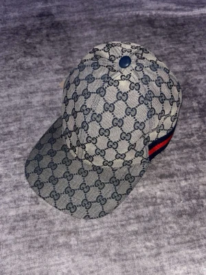 Gucci keps med GG-mönster - Säljer en beige keps från Gucci med klassiskt GG-mönster i mörkgrått. Kepsen har en böjd skärm och justerbar rem baktill. På sidan finns en snygg detalj med röd och blå rand. Materialet är canvas och kepsen har en cool streetstil.