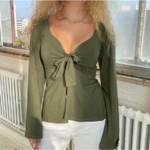 Khakigrön långärmad topp med knyt från designbysi - Trendig olivgrön topp med lång ärm och djup v-ringning. Snygg knytning framtill som ger en feminin touch. Tillverkad i ett mjukt tyg som sitter bekvämt och har en figurnära passform. Den är från märket DesignBySi och är endast använd 2 gånger! 