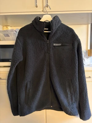 Columbia fleece medium - Svart fleecejacka med dragkedja och hög krage från Columbia. Jackan har en bröstficka med Columbia logga och två sidfickor.  Helt ny.  Nypris 1000kr.  Kvitto finns om det önskas.