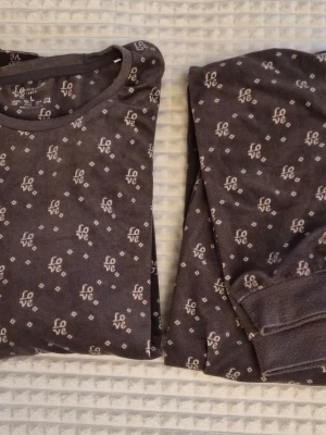 Mjuk brun pyjamas från Primark - Mysig brun pyjamas från Primark med 'love'-mönster över hela setet. Tröjan har rund hals och långa ärmar, byxorna har muddar vid benslut. Perfekt för chillkvällar och att slappa hemma. Materialet är mjukt och skönt mot huden.