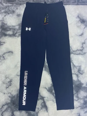 Mörkblå träningsbyxor från Under Armour - Snygga mörkblåa träningsbyxor från Under Armour med reflektiv logga på låret och stort reflektiv tryck längs benet. Byxorna har dragkedja vid benslut och är tillverkade i ett stretchigt funktionsmaterial som passar perfekt till träning eller chill.