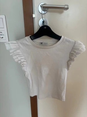 Vit ribbad topp med volangärmar Zara - Supersöt vit ribbad topp från Zara med volangdetaljer på ärmarna. Modellen är croppad och har rund halsringning. Fin till sommaren eller mer festligare tillfälllen🥰 I storlek S 