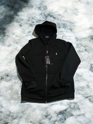 Svart kofta från Polo Ralph Lauren - Snygg svart hoodie med dragkedja från Polo Ralph Lauren. Klassisk design med huva, dragsko och fickor framtill. Liten vit logga broderad på bröstet. Perfekt för en avslappnad och stilren look.