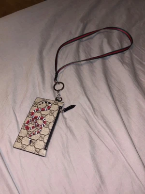 Gucci korthållare med orm och nyckelband - Snygg korthållare från Gucci i beige canvas med klassiskt GG-mönster och en röd orm på framsidan. Baksidan är svart läder med plats för flera kort. Medföljer ett randigt nyckelband i rött och grönt. Perfekt accessoar för dig som vill ha något unikt och trendigt.