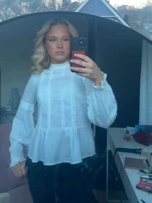 Vit volangblus med hög krage - Säljer en söt vit blus med volanger och hög krage. Blusen har genombrutna detaljer, puffiga långärmar med resår vid ärmslut och en lätt transparent look. Blusen är från Kappahl och passar till alla tillfällen 