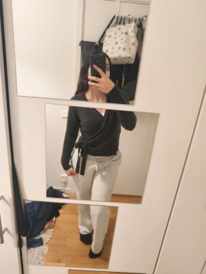 Mörkgrå omlottopp med knytband - Snygg mörkgrå omlottopp med långa ärmar och knytband i midjan. Tröjan har en djup v-ringning och sitter figurnära. Perfekt för en avslappnad men ändå stilren look.