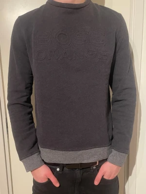 Hugo Boss sweatshirt  - Mörkgrå sweatshirt från HUGO BOSS Orange-kollektionen med broderad logga framtill. Tröjan har rund hals, ribbade muddar och nederkant i ljusare grått. Perfekt för en avslappnad och stilren look. Materialet känns mjukt och bekvämt.