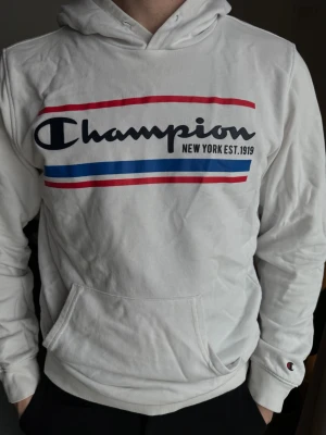 Vit Champion hoodie med tryck - Snygg vit hoodie från Champion med stort tryck på bröstet i svart, rött och blått. Klassisk känguruficka framtill och liten Champion-logga på ärmen. Mjuk bomullskänsla och huva med dragsko. Perfekt för en avslappnad streetwear-look. Utan snören!