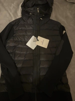 Svart pufferjacka från Moncler - Snygg svart pufferjacka från Moncler med quiltad front och huva. Jackan har stickade ärmar och muddar, dragkedja framtill och Moncler-logga på ärmen. Perfekt för kalla dagar och riktigt trendig look.