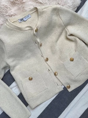 Krämvit kofta från Zara med guldknappar - Stilren krämvit kofta från Zara med rund halsringning och dekorativa guldknappar framtill. Koftan har två fickor fram och är tillverkad i ett mjukt, stickat material.