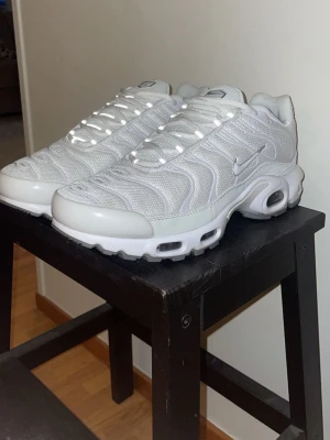 Nike Air Max Plus vita sneakers - Nike Air Max Plus sneakers i helvitt med mesh- och syntetmaterial. Skorna har den ikoniska vågiga designen, synlig Air-dämpning i sulan och små Swoosh-loggor på sidorna. Perfekta för dig som gillar sportig och clean stil.