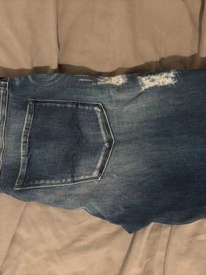 Blå slitna jeans från Replay - Säljer ett par blå jeans från Replay, modell Bronny, med slitningar och en bakficka med dekorativ söm. Jeansen har klassisk femficksdesign och är tillverkade i denim med snygg tvättad look. Perfekta för en avslappnad och trendig stil.