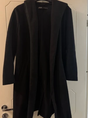 Svart lång kappa  - Svart lång kappa med huva som värmer, ett midjeband medföljer och två fickor med vardera sida. 