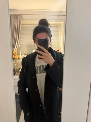 Svart kappa med midjeskärp  - Stilren svart kappa med knappar framtill och tillhörande bälte i midjan. Kappan har klassisk krage och raka ärmar, vilket ger en tidlös look. Passar perfekt till höst och vinter för en snygg och enkel outfit.