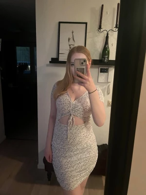 Kort ljus klänning med knyt och cut-out - Säljer en kort ljus klänning med små blommönster, v-ringning och knyt framtill som skapar en snygg cut-out effekt. Klänningen har korta ärmar och figurnära passform, perfekt för sommarens alla tillfällen.