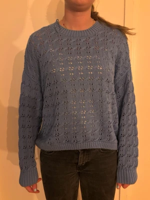 Blå stickad tröja med hålmönster - Säljer en ljusblå stickad tröja med hålmönster och rund hals. Tröjan har långa ärmar och ribbade muddar vid ärmslut och nederkant. Perfekt för dig som gillar en avslappnad men ändå snygg stil.