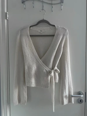 Vit omlottstickad tröja - Säljer en mjuk och fluffig vit stickad omlott-tröja med knytband i sidan. Tröjan har djup V-ringning, långa ärmar och ribbade muddar. Perfekt för lager på lager och ger en avslappnad men ändå stilren look.