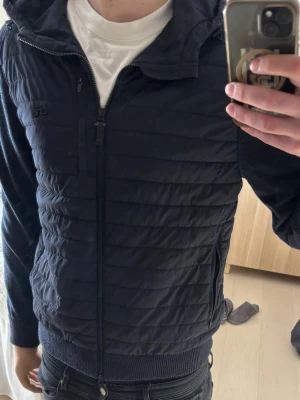 J.Lindeberg Cardigan storlek L - Säljer en mörkblå Cardigan  från J.Lindeberg med quiltad framsida och rygg, hel dragkedja och huva. Jackan har ribbade muddar och sidofickor med dragkedja. Storlek L men passar även M