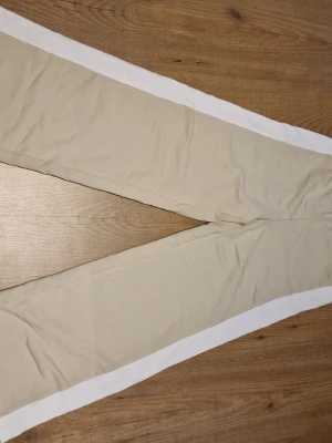 Beige mjukisbyxor med vita revärer - Säljer ett par beige mjukisbyxor med vita revärer längs sidorna. Byxorna har en avslappnad passform och elastisk midja, perfekta för chill dagar. Materialet är mjukt och bekvämt, troligtvis bomull eller polyesterblandning. Perfekt för dig med casual stil. Storlek 152