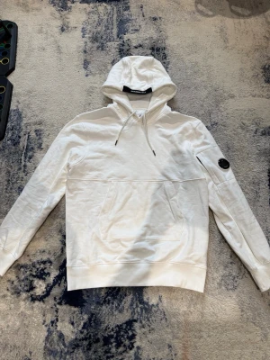 Vit C.P. Company hoodie - Snygg vit hoodie från C.P. Company med klassisk huva och dragsko. Hoodien har en stor magficka och den ikoniska linsen på ena ärmen. Storlek L. Han på bild är 185cm, 75kg
