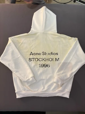 Vit hoodie Acne Studios Stockholm - Säljer en vit hoodie från Acne Studios med tryck på ryggen där det står 'Acne Studios STOCKHOLM 1996'. Hoodien har klassisk huva, ribbade muddar och är gjord i mjukt bomullsmaterial. Perfekt för dig som gillar minimalistisk och stilren design.