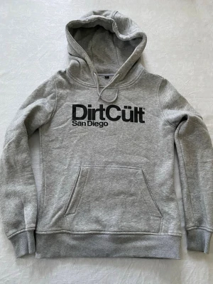 Grå hoodie från Dirt Cult - Snygg grå hoodie från Dirt Cult San Diego med svart tryck på bröstet. Klassisk känguruficka framtill, ribbade muddar och huva med dragsko. Perfekt för en avslappnad streetstyle-look. Mjuk bomullsblandning för extra komfort.