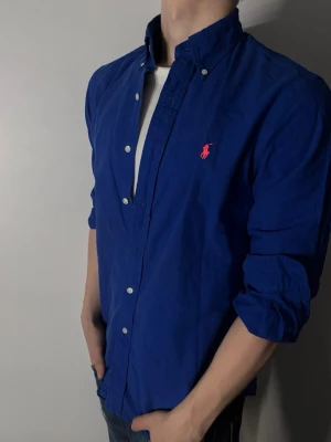 Blå skjorta från Ralph Lauren - Klassisk blå skjorta från Ralph Lauren med knappkrage och den ikoniska röda logotypen broderad på bröstet. Skjortan har långa ärmar och vita knappar framtill. Perfekt för en stilren och avslappnad look.