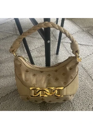 Beige handväska med flätat handtag - Stilren beige handväska med flätat handtag och guldfärgade detaljer. Väskan har ett mönster med små loggor och en stor guldfärgad kedjedekor framtill. Tillverkad i tyg och konstläder, perfekt för dig som gillar trendiga accessoarer. Sliten men supersnygg