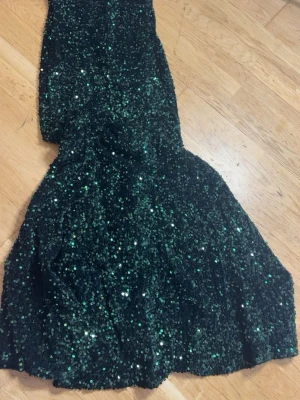 Grön glittrig långklänning - Säljer en fantastisk långklänning i mörkgrönt med massor av glittrande paljetter. Klänningen har en figurnära passform upptill och en utsvängd nederdel som ger en elegant siluett. Perfekt för dig som vill sticka ut på festen! 