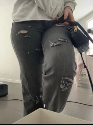 Grå slitna jeans med hål - Säljer ett par grå jeans med flera slitningar och hål på benen. Jeansen har en avslappnad passform och är perfekta för en trendig streetstyle-look. Materialet är klassisk denim och färgen är tvättad grå.