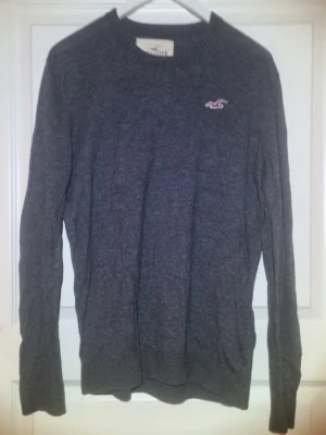 Mörkgrå stickad tröja från Hollister - Mysig mörkgrå stickad tröja från Hollister med rund halsringning och diskret logotyp broderad på bröstet. Tröjan har långa ärmar och ribbstickade muddar vid hals, ärmslut och nederkant. Perfekt för att hålla värmen under kyliga dagar.