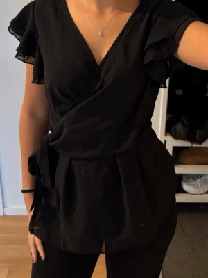 Svart omlottblus med volangärm - Stilren svart omlottblus med volangdetaljer vid ärmarna och knytband i midjan. Blusen har v-ringning och luftig passform, perfekt för en trendig och avslappnad look. Tillverkad i ett lätt tyg som ger ett snyggt fall.