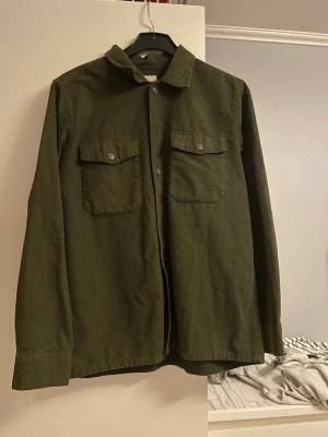 Olivgrön overshirt från Jack & Jones - Stilren olivgrön overshirt från Jack & Jones med två bröstfickor med knappstängning och klassisk krage. Tillverkad i ett mjukt, tjockare tyg som passar perfekt som lager. Knappstängning framtill och raka ärmar ger en clean look.