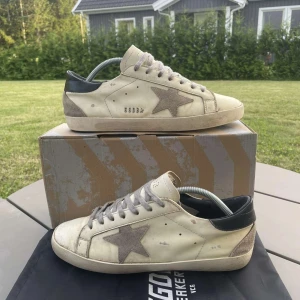 Golden Goose vita sneakers med stjärna - Golden Goose sneakers i vitt skinn med grå mockadetaljer och ikonisk stjärna på sidan. Grå snörning, svart häl och slitna detaljer för en cool, vintage look. Platt sula och rund tå. Perfekta för dig som gillar streetstyle och unika skor. Storlek 44
