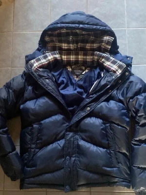 Marinblå pufferjacka från Tommy Hilfiger - Snygg marinblå pufferjacka från Tommy Hilfiger med avtagbar huva och rutigt innerfoder. Jackan har dragkedja och knappar framtill, två sidofickor och en liten logga på bröstet. Perfekt för kalla dagar och riktigt skön att bära.