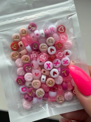 Mixade bokstavspärlor i rosa toner - En påse med mixade bokstavspärlor i olika nyanser av rosa, vitt och guld. Pärlorna har bokstäver och hjärtan i olika former och storlekar, perfekta för att skapa personliga smycken som armband eller halsband.