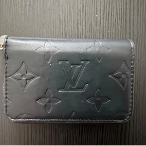 Plånbok  - Ny, liten svart plånbok med silvrig dragkedja och mönster på. Fack för kort mm.. Kommer med box också, om man vill ha. Ca 10.5 cm och 7 cm hög.