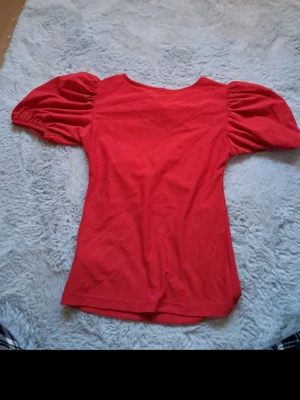 Röd topp med puffärmar från Gina Tricot - Topp från Gina Tricot med puffiga korta ärmar och rund halsringning. Röd blus ifrån ginatricot i strolek xxs. Blusen har ett mellanrum i mitten som jag försökt visa på bilderna. Den är i mycket fint skick.💕